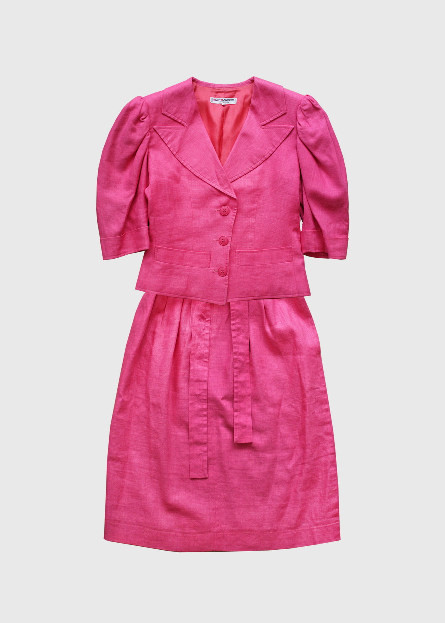 A fabulous, bright pink linen 2 piece set from 1985 Yves Saint Laurent. The dress comes includes a matching belt, knotted to one side and has button details at the back. The jacket has structured shoulders and puff sleeves and featured three matching round pink buttons and two pockets on its front bodice.
