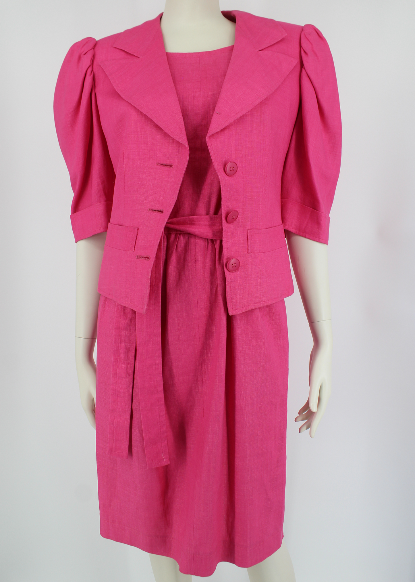 A fabulous, bright pink linen 2 piece set from 1985 Yves Saint Laurent. The dress comes includes a matching belt, knotted to one side and has button details at the back. The jacket has structured shoulders and puff sleeves and featured three matching round pink buttons and two pockets on its front bodice.