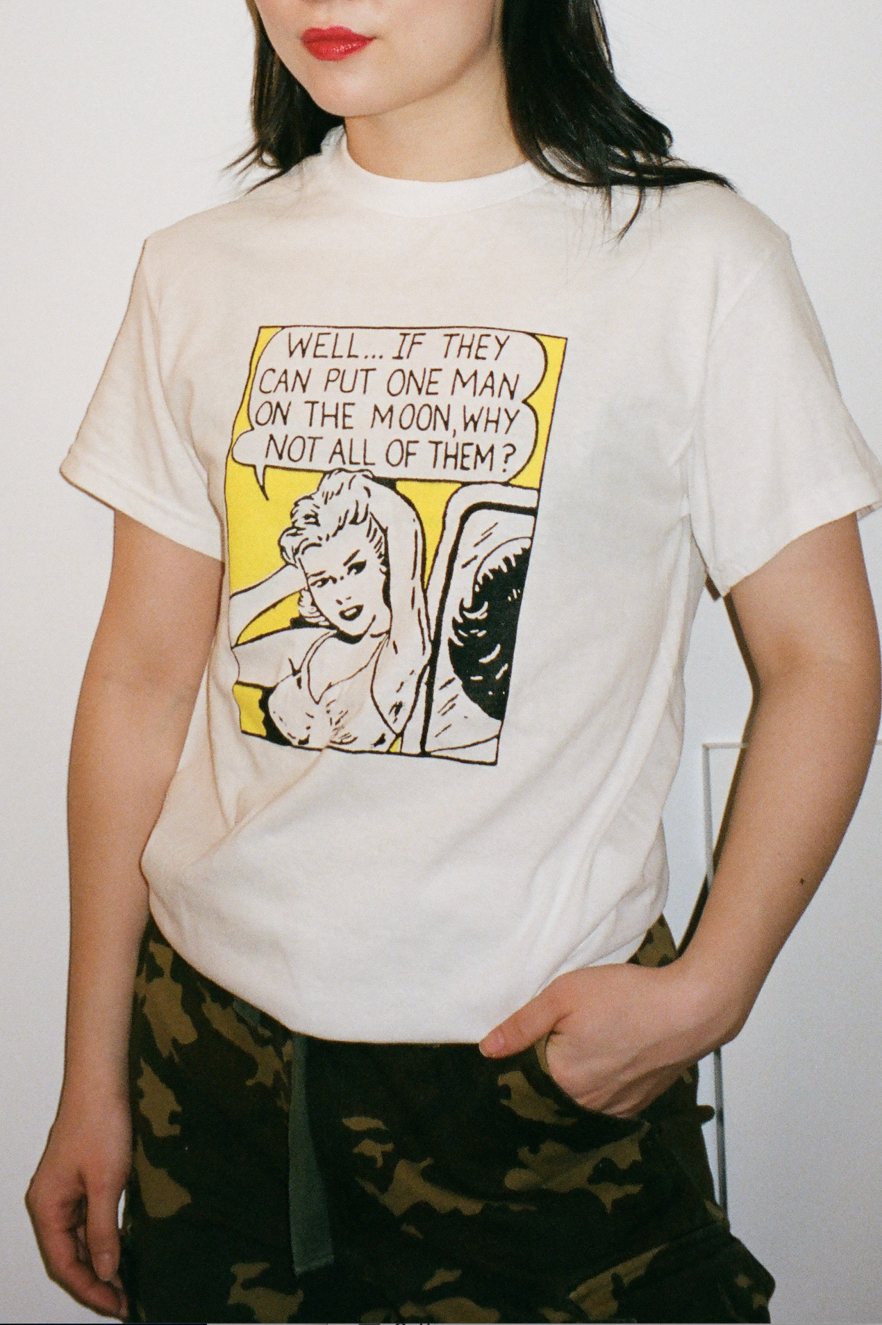 A white cotton t-shirt with a black, white, and yellow square image of a woman saying "Well... if they can put one man on the moon, why not all of them?" centers on the chest of the shirt.