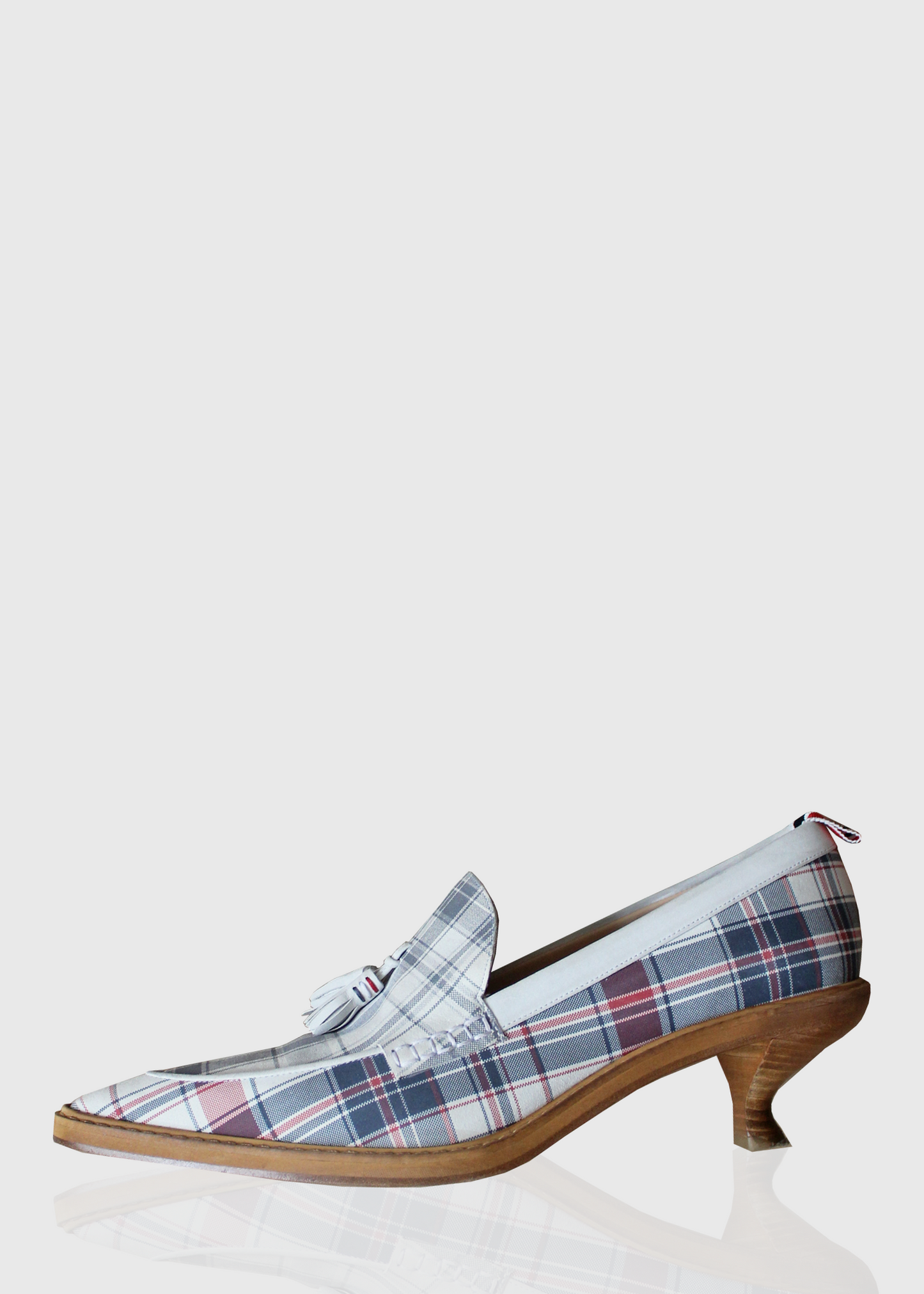 A side view of the Thom Browne shoes that feature a very pointed toe, tassels, and a stacked, curved wooden kitten heel. Rendered in signature blue and white Thom Browne plaid and a red, white, and blue stripped lining.